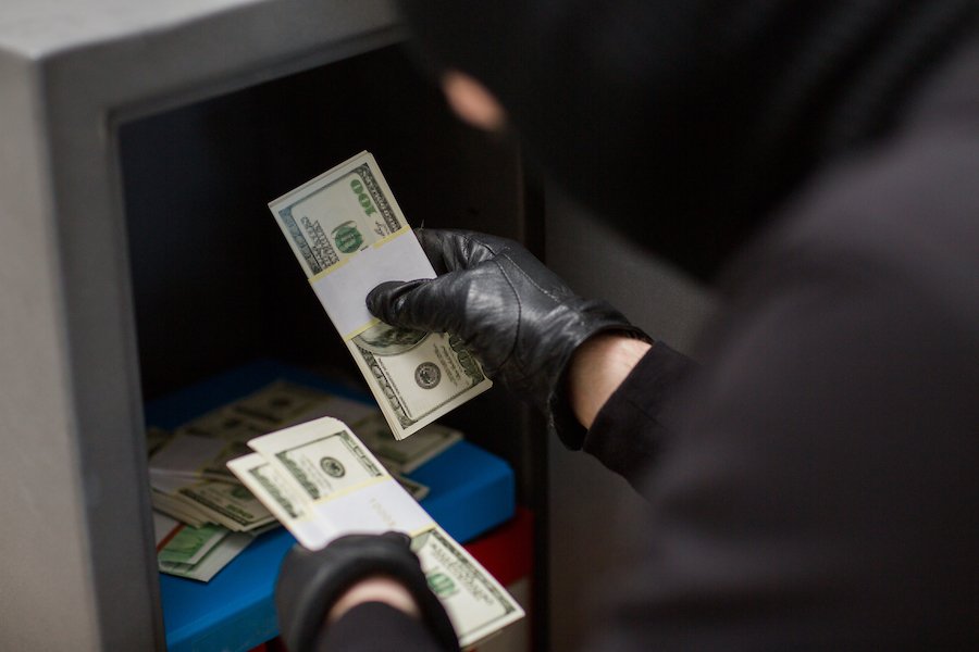 Closeup of masked and gloved man stealing wads of cash from a safe he broke into in violation of NRS 205.220.