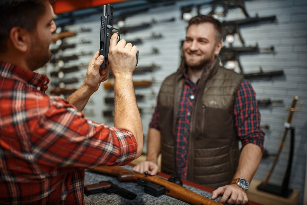 Gun shop worker showing a customer a revolver with an array of rifles on the back wall
