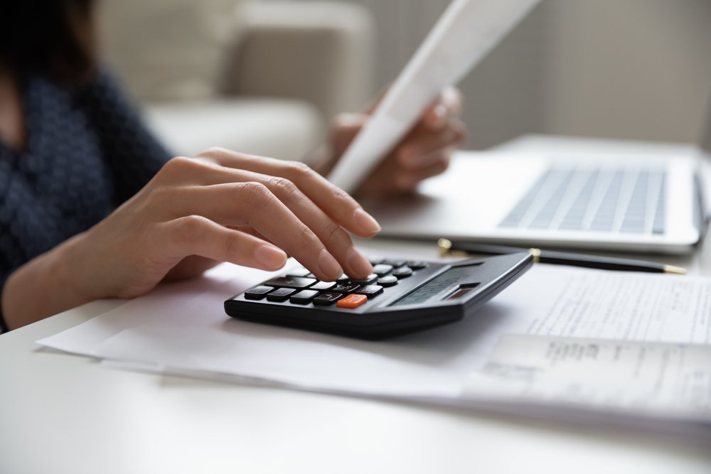 Closeup of a woman's hand on a calculator near a laptop and she holds insurance documents in the other hand