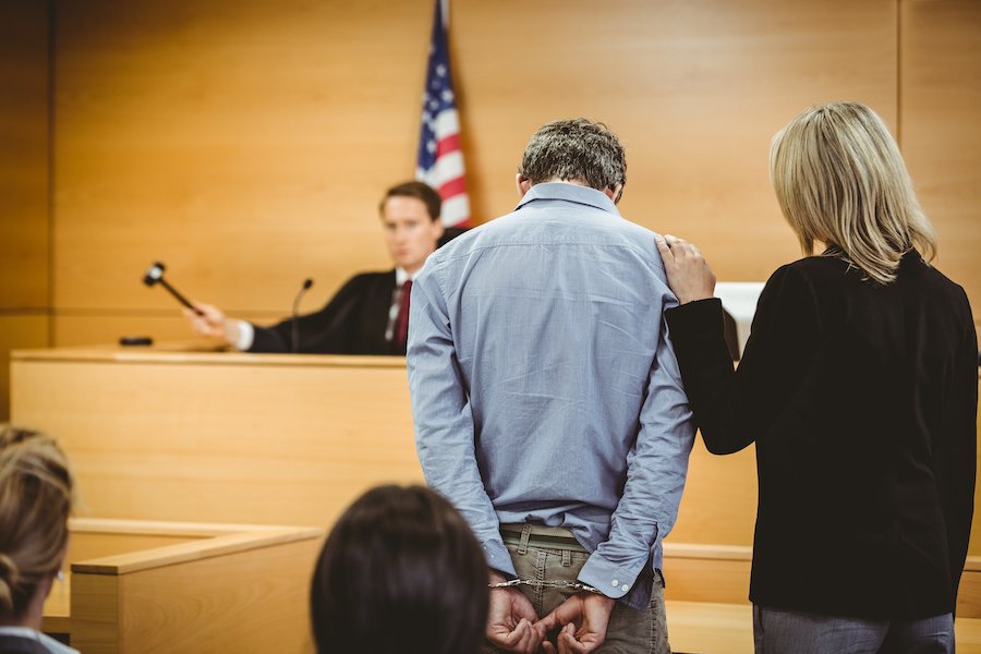 Courtroom where a judge in front of an American flag with a gavel sentences a defendant to a consecutive sentence. The defendant is handcuffed and bowing his head while his attorney comforts him.