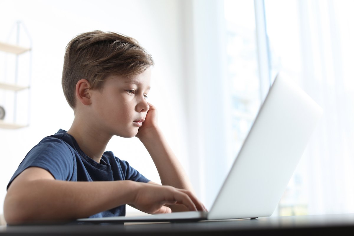 Niño pequeño mirando su laptop en un dormitorio mientras es contactado por un extraño que intenta engañarlo