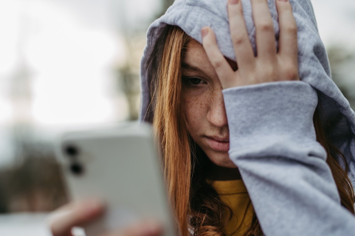 Teen girl in a hoodie looking distraught towards her cell phone