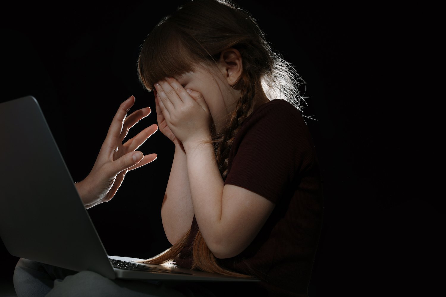 Extraño alcanzando a un niño asustado a través de la pantalla de una laptop contra un fondo oscuro.