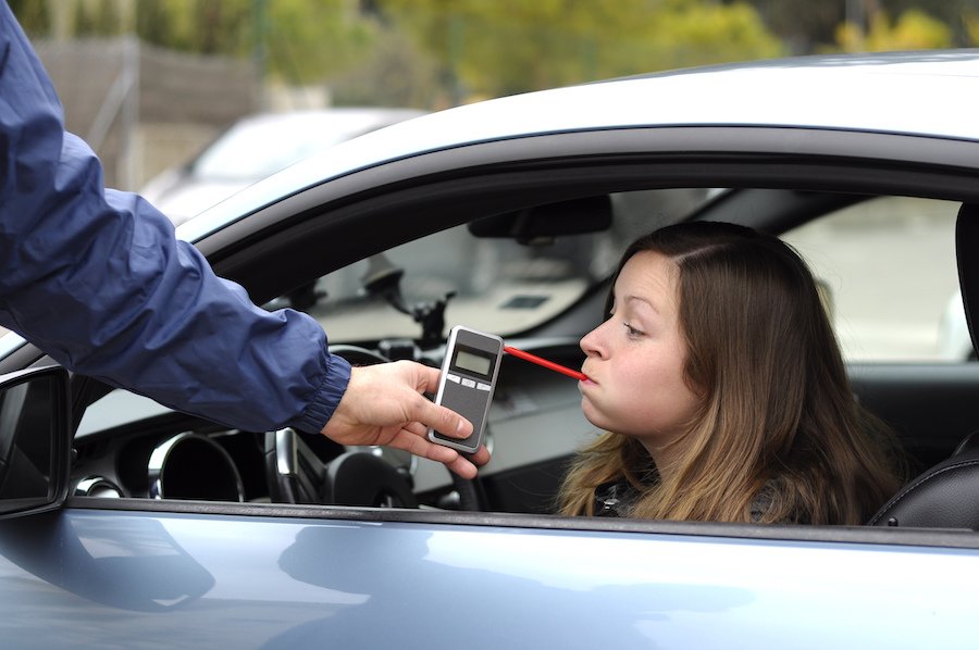 Woman driver parked in a sedan taking a preliminary breath test by blowing into a breathalyzer held by a police officer 