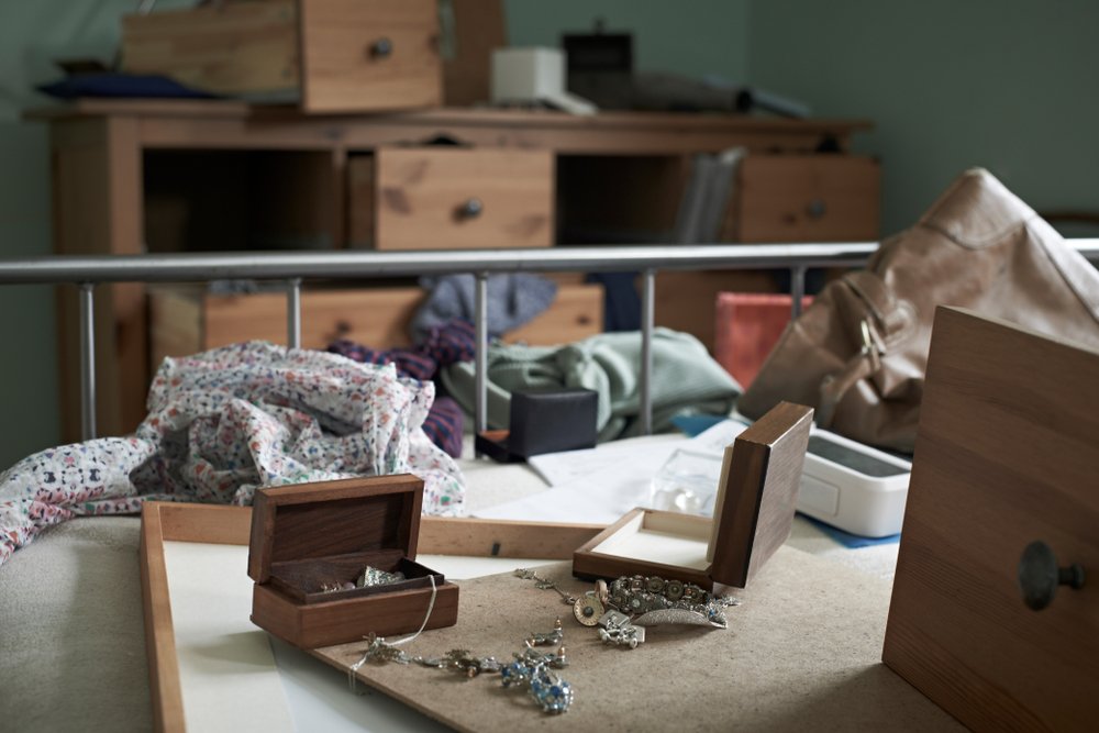 A ransacked bedroom with various pieces of jewelry strewn on the bed and drawers from the bureau pulled out.