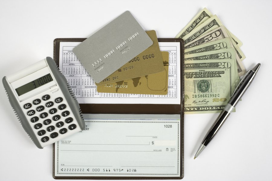 Calculator, three credit cards, a checkbook, cash, and a pen spread out on a white desk