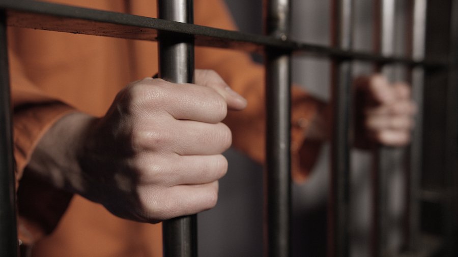 Prisoner's hand gripping prison bars, and the prisoner is wearing an orange jumpsuit