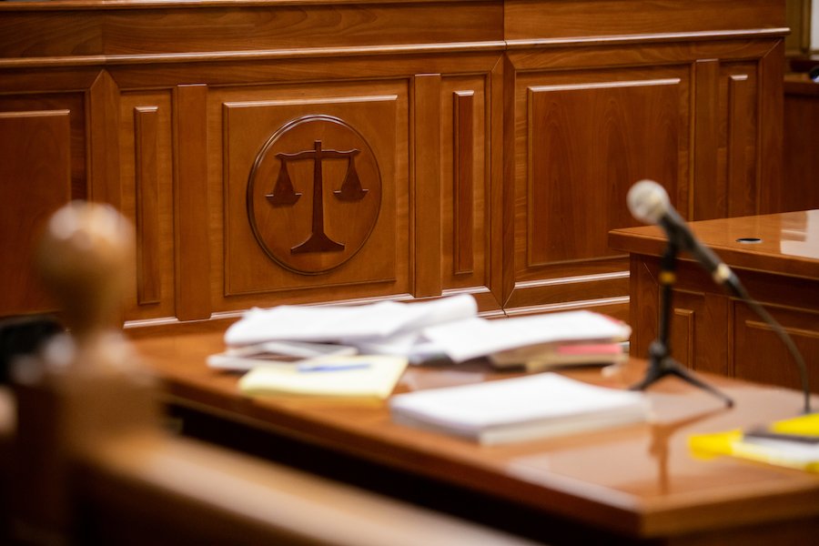 Counsel's desk in a courtroom, covered in papers and with a scale of justice sign engraved into the wood panneling of the wall