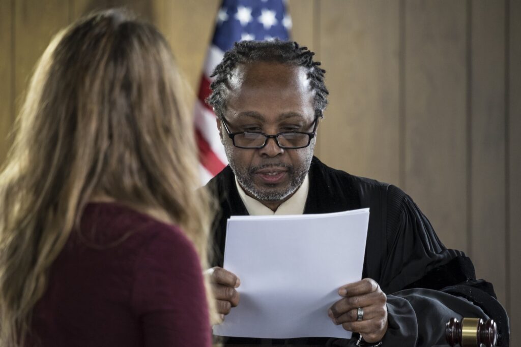 Judge in black robe in front of an American flag speaking to defendant in his courtroom