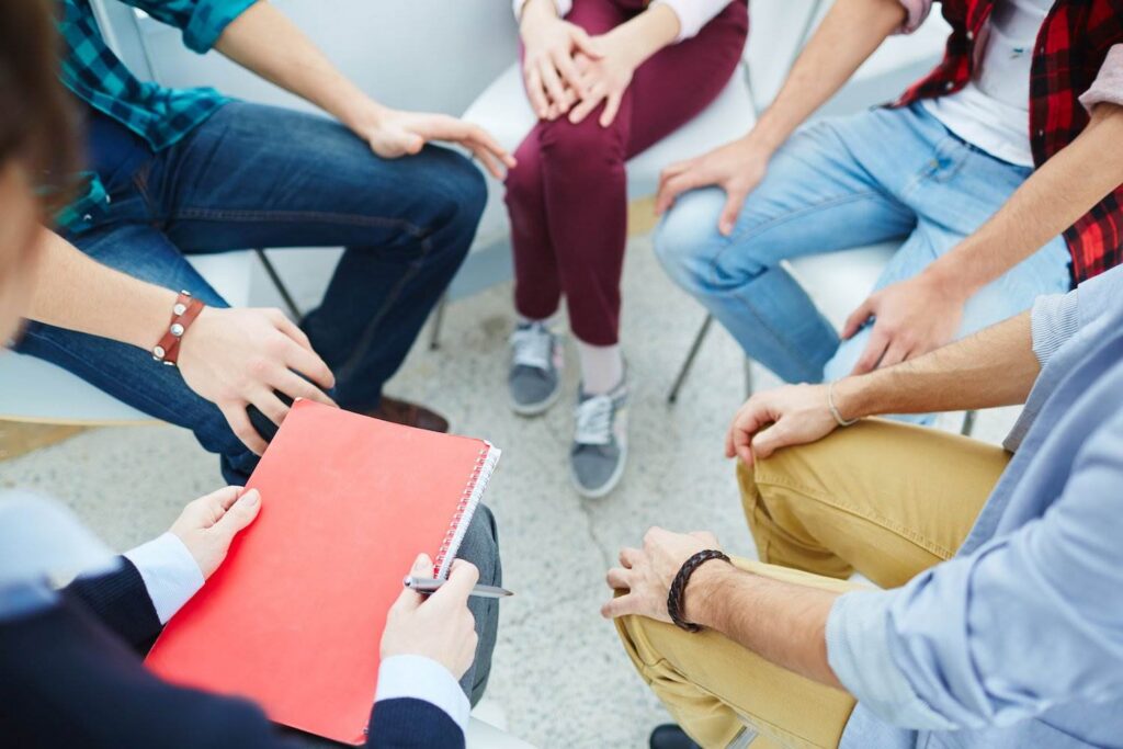 Group counseling session with five people sitting in a circle, and the therapist is holding a notebook