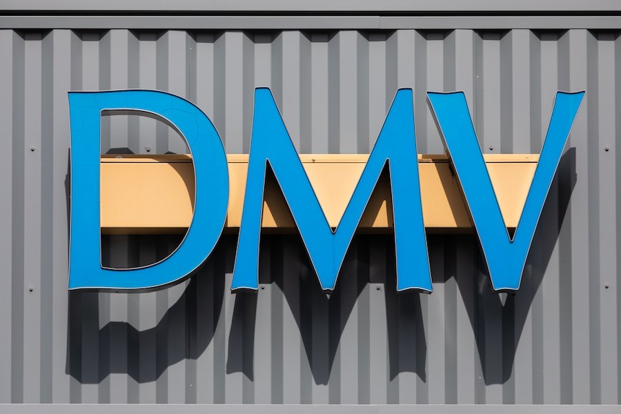 Blue DMV sign on the outside of a DMV office with a grey aluminum siding wall