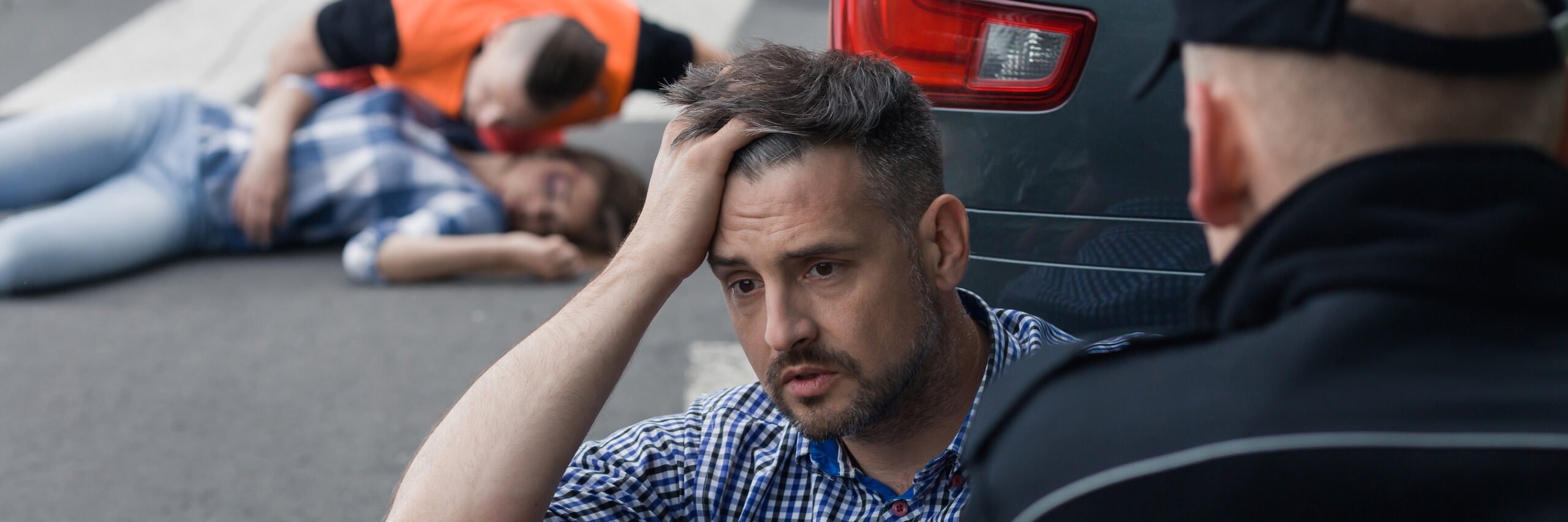 Dazed looking driver being questioned by police following an accident he caused with the victim lying on the ground in the background with an officer examining her