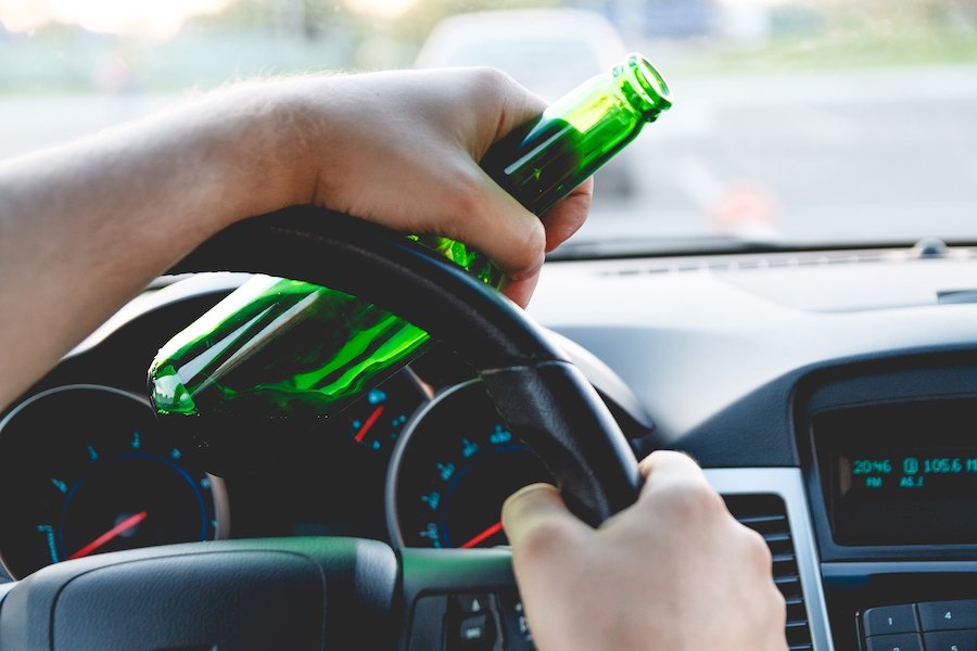 Daytime close-up of a driver's hands with one on the steering wheel and the other holding a beer bottle 