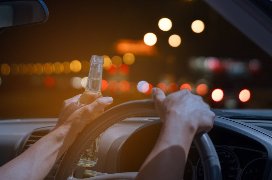 Close up of hands on steering wheel with one holding a beer bottle with a view of the blurred city lights in the distance through the windshield