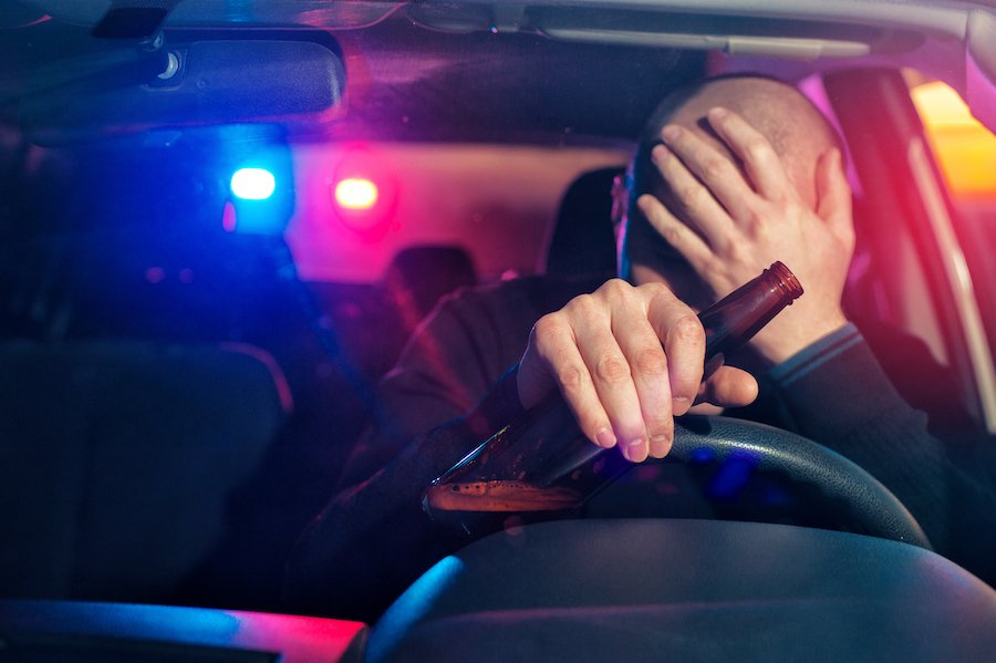 Upset driver holding his head with one hand and the wheel and a beer bottle in the other with police lights visible through the rear windshield signalling for him to pull over