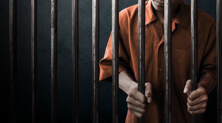 Inmate in orange jumpsuit gripping his prison bars against a gray background