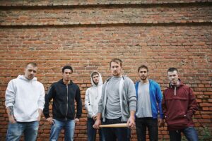 Group of six angry-looking gang members in hoodies against a brick wall, and one is holding a bat