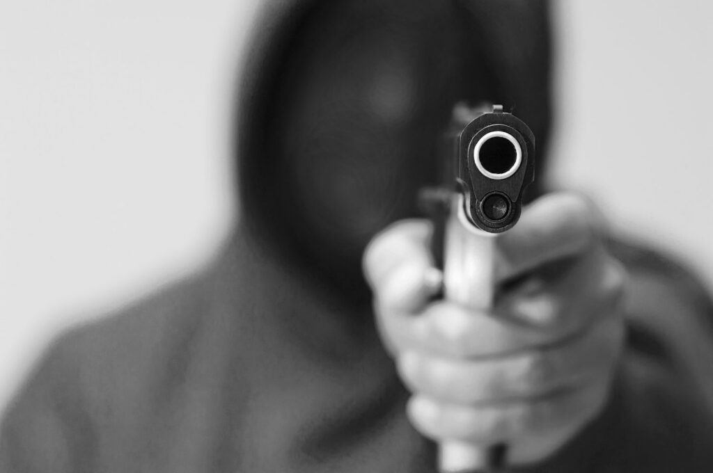 Black and white image of a hooded man pointing gun to the camera, and he is about to violate CRS 18-3-102.