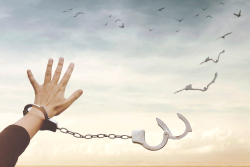 handcuffs against a grey sky with one the cuffs holding a hand while the other is cracked open, mirroring the birds in the background flying to freedom