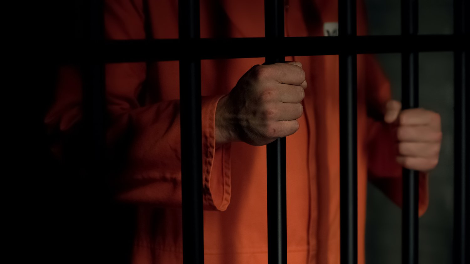 Inmate in orange jumpsuit gripping his prison cell bars