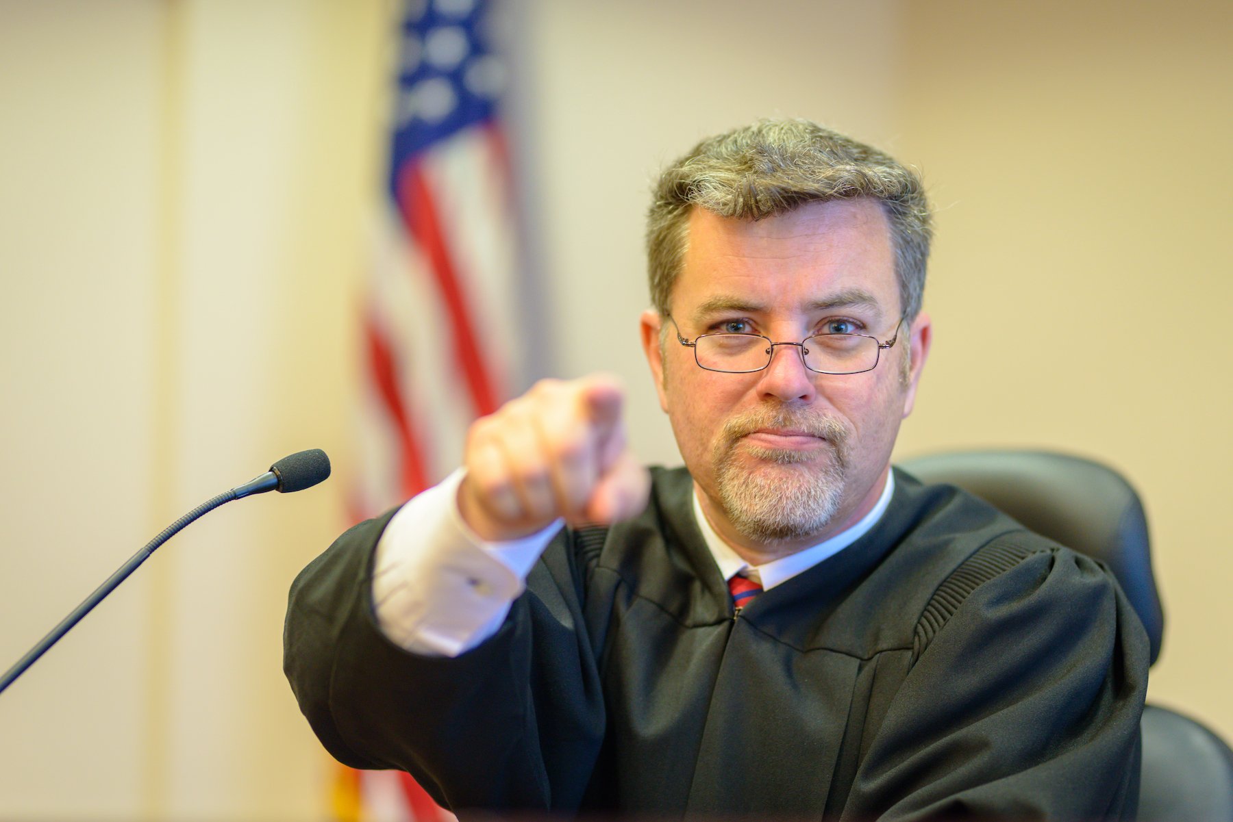 Judge at the bench pointing at the camera with a flag in the background