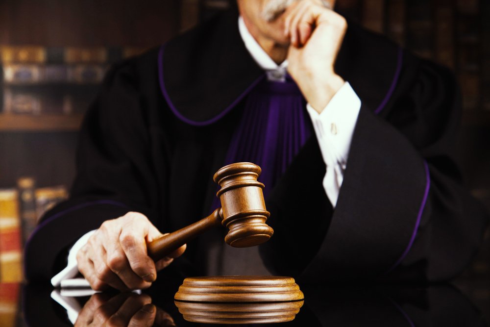 Judge in velvet robe holding his chin with one hand and holding a gavel in the other