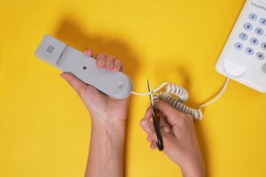Hand with scissors snipping line connecting handset to telephone in violation of Colorado law.